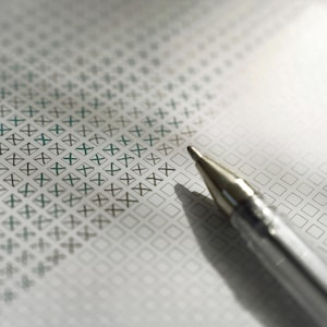 Pu&ograve; includere: Primo piano di una penna d'argento appoggiata su un modulo con una griglia di caselle, alcune contrassegnate con una 'X' verde. La carta ha un motivo sottile. L'ombra della penna &egrave; visibile.