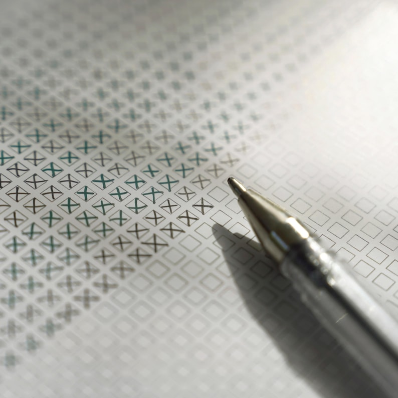 Pu&ograve; includere: Primo piano di una penna d'argento appoggiata su un modulo con una griglia di caselle, alcune contrassegnate con una 'X' verde. La carta ha un motivo sottile. L'ombra della penna &egrave; visibile.