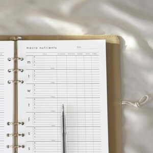 May include: A beige planner with a macro nutrients chart and a silver pen. The chart includes sections for meal, calories, protein, carbs, fats, and fiber. The planner is open to a page with the words "macro nutrients" at the top.