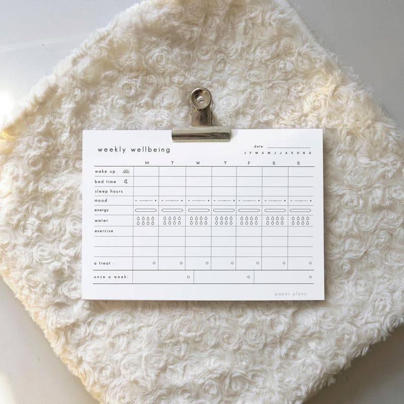 May include: A white paper planner with the words "weekly wellbeing" at the top. The planner has sections for tracking wake up, bed time, sleep hours, mood, energy, water, and exercise. A silver clip holds the planner.
