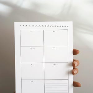 May include: A white weekly planner with a minimalist design. The planner features a grid layout with boxes for each day of the week, labelled with the days of the week. The top of the planner has the months of the year and the days of the month.