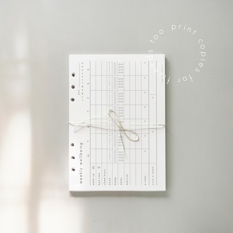 May include: A white weekly wellbeing planner with a grid layout, tied with twine. The planner includes sections for tracking wake up, bed time, sleep hours, mood, energy, water, exercise, and treats. Text on the planner reads "weekly wellbeing" and "too print copies for".