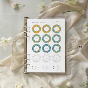 May include: A clear plastic six-ring binder containing a 2027 weather tracker insert. The page features twelve circular charts for each month, with color-coded sections for weather conditions including sun, clouds, and rain, labeled paper plans.