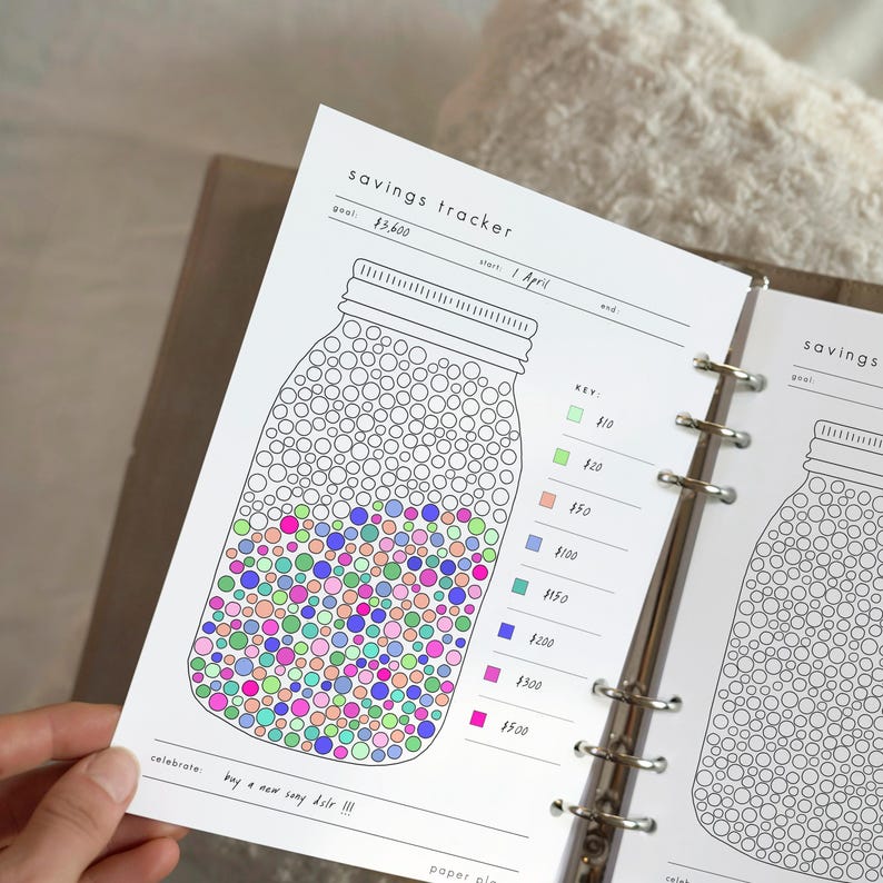 May include: A savings tracker page with a mason jar design. The jar is filled with colorful dots representing savings milestones. The text "savings tracker" and a goal amount are at the top. The text "buy a new sony dslr !!!" is at the bottom.