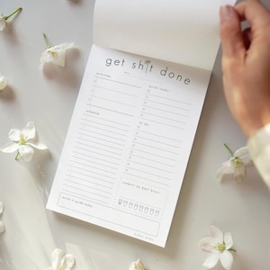 Può includere: Un blocco note per pianificazione in carta bianca con testo e linee nere. L'intestazione recita get shit done. Le sezioni includono priorities, quick tasks, schedule, to do, support my best brain con otto icone di tazze e words to guide today.