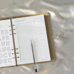 May include: A beige ring-bound planner open to a page titled energy levels with a grid for days one through thirty and months January through December. The page includes a key section with checkboxes and the text paper plans. A black pen rests on the page.