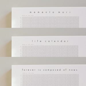 Pu&ograve; includere: Tre calendari di carta bianca con design minimalisti. Il calendario in alto recita "memento mori", quello centrale "life calendar" e quello in basso "forever is composed of nows". Ogni calendario ha una griglia per tenere traccia delle settimane dell'anno.