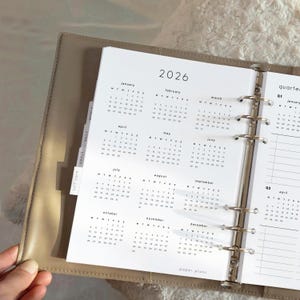 May include: Open planner with a 2026 calendar page. The calendar is printed in black on white paper, with monthly layouts. The planner is in a beige leather-like binder. Tabs labeled "self care", "goals", and "daily" are visible.