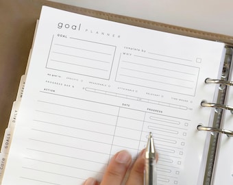 May include: A white goal planner page with the words "goal PLANNER" at the top. The page has sections for goals, actions, and progress tracking. A silver pen is held over the page. The planner is inside a tan binder.