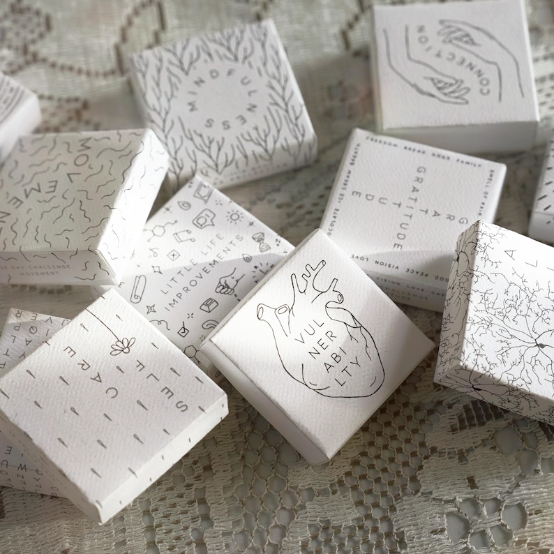 May include: A collection of small, white square boxes with black line art and text. The boxes feature various designs, including a heart, hands, and the words "Gratitude" and "Mindfulness". The overall aesthetic is minimalist and modern.