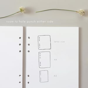 May include: White paper with a sketched design of three different-sized rectangles, labeled "letter size", "A4", and "A5". The text "room to hole punch either side" is above the paper. Two sprigs of white flowers are on the top.