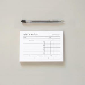 Op de afbeelding: Een wit rechthoekig workout-plannerblok met zwarte tekst en lijnen, voorzien van secties voor datum, focus, tijd, oefening, sets, herhalingen, cardio, afstand, hartslag en calorieën, vergezeld van een doorzichtige balpen.