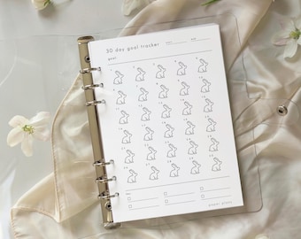May include: A white 30-day goal tracker planner with a clear plastic cover. The planner features a grid of bunny illustrations for each day, with the text "30 day goal tracker" at the top. The planner is held together with a silver binder.