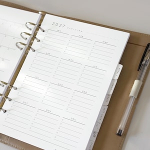 May include: A tan ring-bound planner open to a 2027 overview page with twelve monthly sections, each featuring horizontal lines and small circles for checklists. The planner includes white tabbed dividers labeled weekly, daily, goals, and self care.
