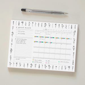 May include: A white monthly planner with floral illustrations and the text "a good month." The planner includes sections for tasks, goals, and a calendar grid. A clear pen rests at the top. The planner is designed for organization.