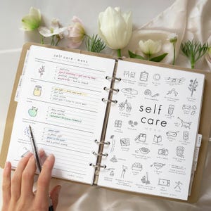 May include: Open planner with a "self care menu" on the pages. The left page has handwritten notes and illustrations. The right page has a variety of small black and white illustrations. The planner is open on a white surface with flowers.