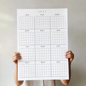 May include: A white wall calendar for the year 2027 featuring a grid layout for all twelve months. Each month is labeled with its name and includes a grid for days of the week. The text 2027 is printed at the top and paper plans at the bottom.