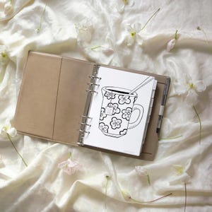 May include: A beige leather six-ring binder open to a page featuring a black and white line drawing of a floral patterned mug with a tea bag tag that reads "it's the little things". A black pen is held in the binder's side loop.