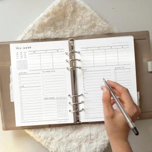 May include: A weekly planner with sections for tasks, meals, and a grocery list. The planner is open to a page with the text "this week". A hand holds a pen over the page, ready to write. The planner is in a neutral-toned binder.