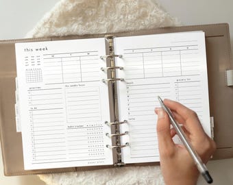 May include: Open planner with weekly layout, including sections for priorities, to-do list, and habit tracker. The planner is open to a page with the text "this week". A hand holds a pen over the page.