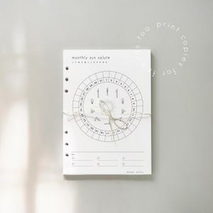 May include: A white planner page with a circular diagram for tracking yoga poses, labeled "monthly sun salute." The diagram is surrounded by numbers and illustrations of yoga positions. The page is tied with twine and has the text "paper plans."