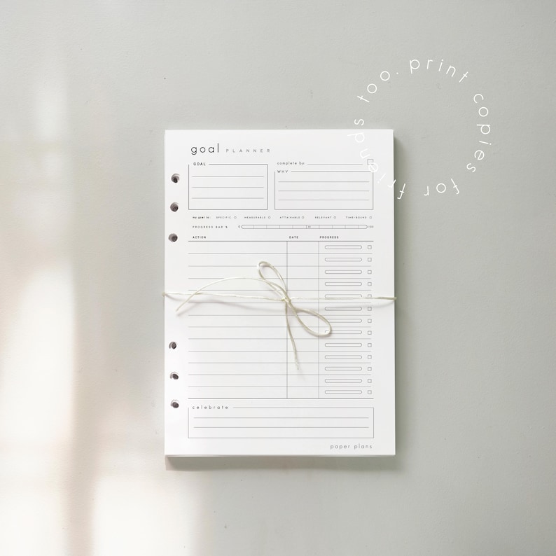 May include: A white goal planner with the text "goal planner" at the top. The planner has sections for goals, actions, dates, and progress. It is bound with a white string.