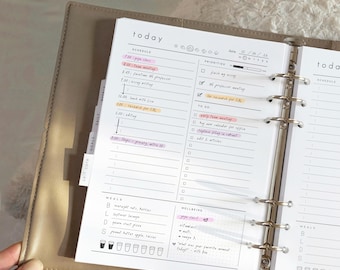 May include: A beige ring-bound planner open to a daily schedule page. The white paper features sections for today's schedule, priorities, to-do lists, meals, and wellbeing. Handwritten entries are highlighted in pink and orange ink.