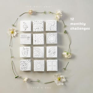 May include: A collection of twelve white square cards, each with a unique black and white design and text, arranged with flowers. The cards are labeled with phrases like "Impact" and "Gratitude". The text "the best year", "12 monthly challenges", and "1 card a day" are also visible.