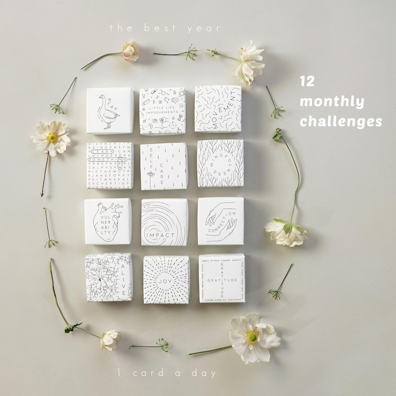 May include: A collection of twelve white square cards, each with a unique black and white design and text, arranged with flowers. The cards are labeled with phrases like "Impact" and "Gratitude". The text "the best year", "12 monthly challenges", and "1 card a day" are also visible.
