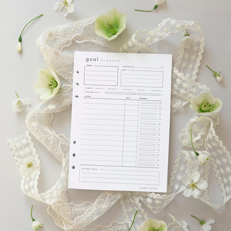 May include: A white goal planner with a minimalist design, featuring sections for goals, actions, and progress tracking. The planner is surrounded by white lace ribbon and delicate white and green flowers. The text "GOAL PLANNER" is visible at the top.