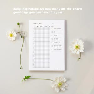 K&ouml;nnte beinhalten: Ein wei&szlig;er Notizblock mit einem Raster zur t&auml;glichen Verfolgung, mit dem Text "rate my day" und "daily inspiration: see how many off-the-charts good days you can have this year!". Wei&szlig;e Blumen umgeben den Notizblock.