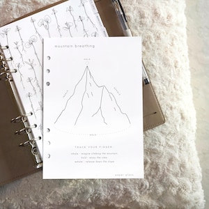 Könnte beinhalten: Eine weiße Planereinlage mit einer schwarzen Strichzeichnung eines Berges und dem Text mountain breathing, inhale, exhale, hold, trace your finger sowie paper plans. Die Seite enthält Anleitungen für eine Atemübung.