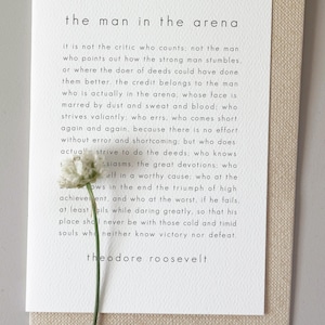 May include: A white rectangular card featuring the printed text of The Man in the Arena by Theodore Roosevelt in a black sans-serif font. A single small white flower with a thin green stem is positioned over the lower left portion of the text.