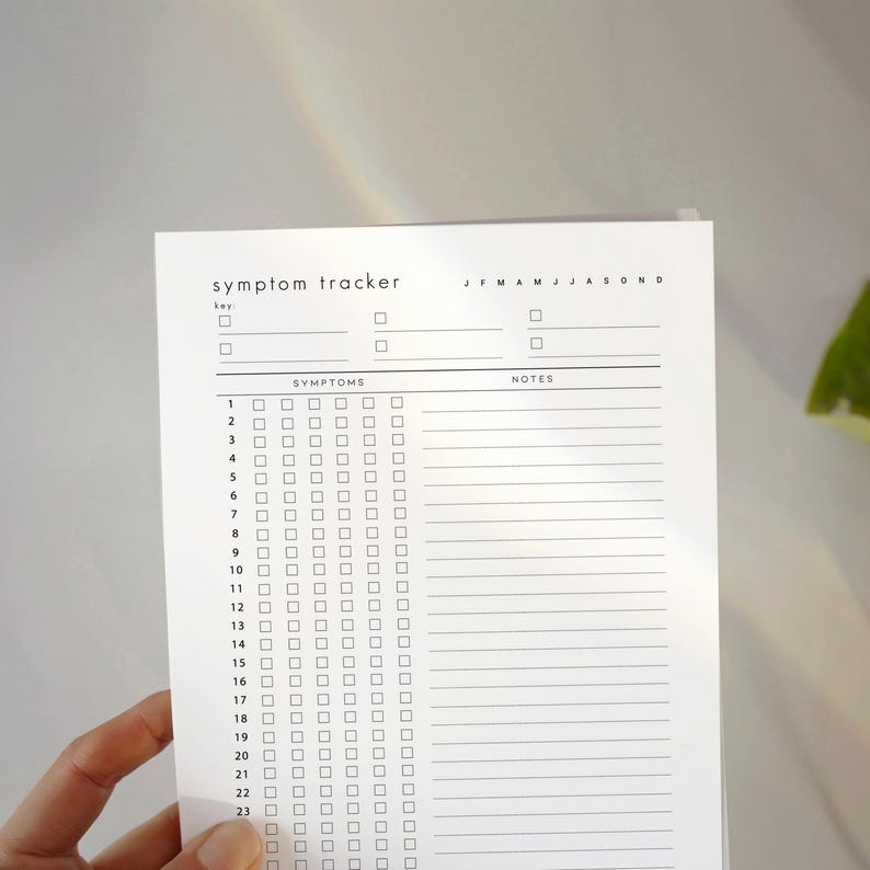 May include: A white symptom tracker sheet with the title "symptom tracker" at the top. The sheet has a key, a section for symptoms, and a notes section. The months of the year are listed across the top. The sheet is held by a hand.