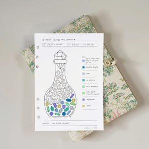 Op de afbeelding: Een witte papieren planner met de tekst "prioritizing my peace" en een illustratie van een pot gevuld met kleurrijke edelstenen. De planner bevat een sleutel met zelfzorgdoelen en ligt op een bloemmotief.