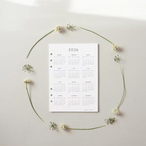 May include: A white 2026 calendar with a minimalist design, featuring monthly layouts. The calendar is surrounded by a delicate arrangement of small white flowers and green stems, creating a natural frame. The text "paper plans" is at the bottom.