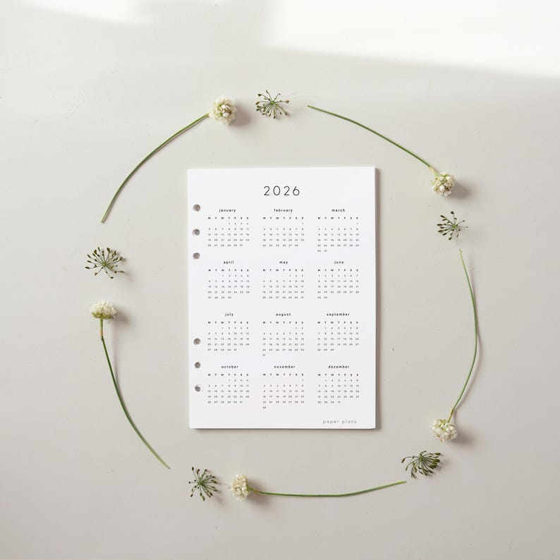 May include: A white 2026 calendar with a minimalist design, featuring monthly layouts. The calendar is surrounded by a delicate arrangement of small white flowers and green stems, creating a natural frame. The text "paper plans" is at the bottom.