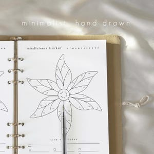 Puede incluir: Un rastreador de atenci&oacute;n plena minimalista dibujado a mano con un dise&ntilde;o floral. La ilustraci&oacute;n en blanco y negro presenta p&eacute;talos numerados y el texto "mindfulness tracker" y "JFMAMJJASOND". Un bol&iacute;grafo apunta al centro de la flor, dentro de una carpeta beige.