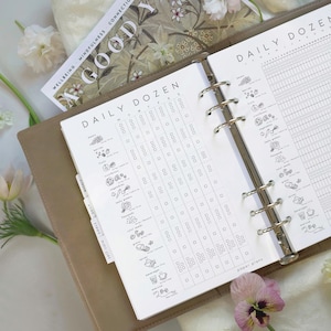 May include: Open planner with "DAILY DOZEN" checklist pages. The pages are white with black text and illustrations. A magazine with the word "GOOD" is visible. The planner is tan and open to reveal the pages. Flowers and greenery are in the background.