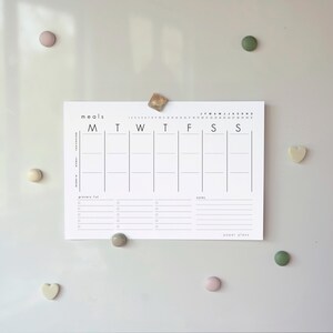 Può includere: Un pianificatore di pasti bianco con testo e linee nere, tenuto in posizione da diversi magneti colorati. Il calendario include sezioni per ogni giorno della settimana, una lista della spesa e note. Il testo "meals" e "paper plans" è visibile.