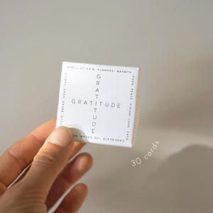May include: A small white box of gratitude cards held in a hand. The word "GRATITUDE" is printed in a cross shape on the front. The box also has the words "30 cards" printed on it. The box has words like "flowers" and "cats" printed on the sides.