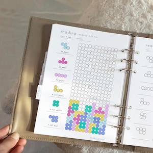 May include: An open planner with a reading tracker titled "BUBBLE TETRIS." The page has a grid of circles to track reading progress, with colourful Tetris-like shapes filling in as goals are met. The planner includes tabs for self-care, goals, and daily tasks.
