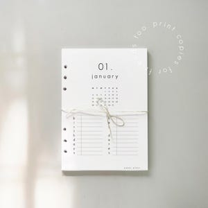 May include: A white paper planner with the date "01. January" printed at the top. The planner includes a calendar, lines for birthdays, and the text "paper plans". It is tied with a string.