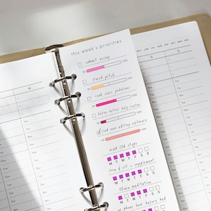May include: A white planner insert with a metal six-ring binder mechanism. The page features a 'this week's priorities' section with checkboxes, progress bars in pink and orange, and habit trackers for steps, vitamins, meditation, and phone usage.