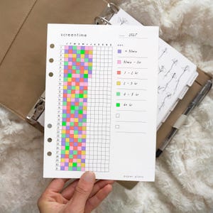 Op de afbeelding: Een wit papieren planner-inlegvel voor 2027 met de titel screentime en een raster voor het bijhouden van dagelijkse gewoontes. Het bevat een kleurgecodeerde legenda met paarse, roze, oranje, gele en groene vierkanten voor verschillende tijdsduren.