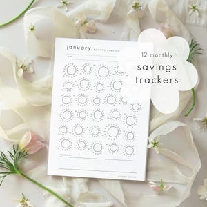 May include: A white paper savings tracker sheet titled January Savings Tracker with 12 monthly savings trackers text. The page features a goal section, a grid of thirty-one sun-shaped icons for daily tracking, and a celebrate section at the bottom.