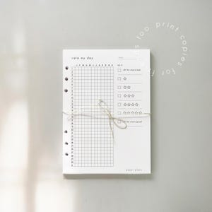K&ouml;nnte beinhalten: Ein wei&szlig;er Papierplaner mit dem Text "rate my day" oben. Der Planer hat ein Raster zur t&auml;glichen Verfolgung und einen Schl&uuml;ssel zur Bewertung. Er ist mit einer Schnur gebunden und enth&auml;lt den Text "paper plans".
