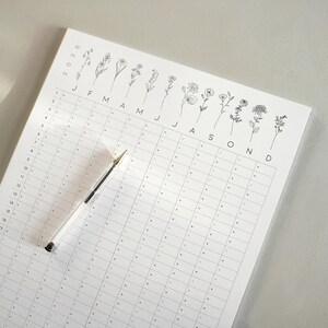 May include: A white 2026 year planner featuring a floral design. The planner has a grid layout with dotted lines and a clear pen. The floral design at the top includes delicate line drawings of various flowers. The year and months are labelled.