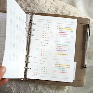 May include: A tan ring-bound planner open to a 2027 quarterly plan page. The page features calendars for January, February, and March with handwritten notes and colourful highlights. A clear plastic pen is held in a loop on the side of the cover.