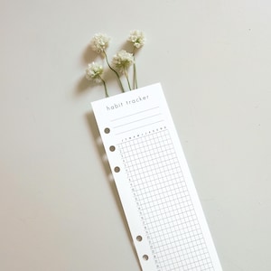 Puede incluir: Una hoja de seguimiento de hábitos en papel blanco con texto y líneas de cuadrícula en negro. La parte superior muestra el texto habit tracker, seguido de tres líneas en blanco. Una cuadrícula muestra los meses J a D y los días 1 a 31. Cinco agujeros.